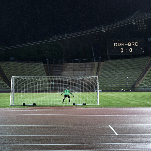 Die Fotografie zeigt das Innere des Olympiastadions bei Nacht. Zu sehen ist der Künstler Massimo Furlan im grün-schwarzen Fußballtrikot in Rückenansicht, der in einem Tor steht. Vor ihm erstreckt sich der grüne Fußballrasen, er ist jedoch der einzige Spieler auf dem Platz. Auf einer Anzeigetafel hoch über den Zuschauerrängen erscheint der Spielstand dieses fiktiven Fußballspiels: "DDR gegen BRD – 0 zu 0".
