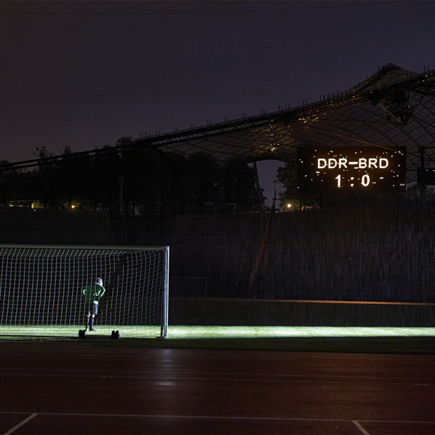 Die Fotografie zeigt das Innere des Olympiastadions bei Nacht. Zu sehen ist der Künstler Massimo Furlan im grün-schwarzen Fußballtrikot in Rückenansicht, der in einem Tor steht. Vor ihm erstreckt sich der grüne Fußballrasen, er ist jedoch der einzige Spieler auf dem Platz. Nur der Spot eines Flutlichts auf dem Rasen erhellt das sonst dunkle Stadion. Auf einer Anzeigetafel hoch über den Zuschauerrängen erscheint der Spielstand dieses fiktiven Fußballspiels: "DDR gegen BRD – 1 zu 0".