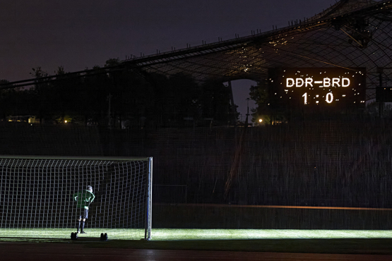 Die Fotografie zeigt das Innere des Olympiastadions bei Nacht. Zu sehen ist der Künstler Massimo Furlan im grün-schwarzen Fußballtrikot in Rückenansicht, der in einem Tor steht. Vor ihm erstreckt sich der grüne Fußballrasen, er ist jedoch der einzige Spieler auf dem Platz. Nur der Spot eines Flutlichts auf dem Rasen erhellt das sonst dunkle Stadion. Auf einer Anzeigetafel hoch über den Zuschauerrängen erscheint der Spielstand dieses fiktiven Fußballspiels: "DDR gegen BRD – 1 zu 0".