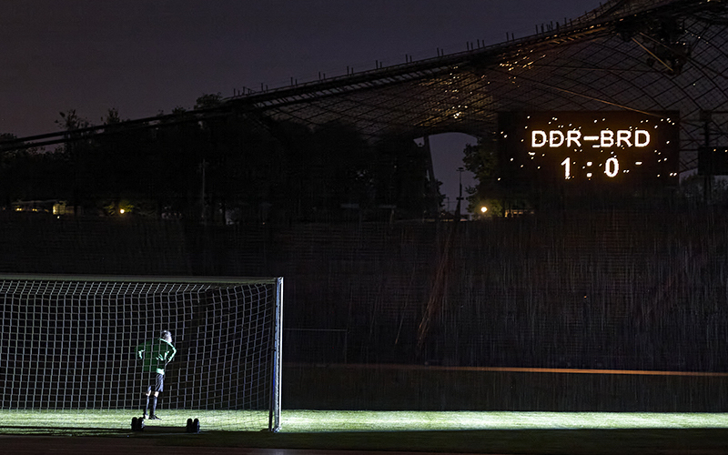 Die Fotografie zeigt das Innere des Olympiastadions bei Nacht. Zu sehen ist der Künstler Massimo Furlan im grün-schwarzen Fußballtrikot in Rückenansicht, der in einem Tor steht. Vor ihm erstreckt sich der grüne Fußballrasen, er ist jedoch der einzige Spieler auf dem Platz. Nur der Spot eines Flutlichts auf dem Rasen erhellt das sonst dunkle Stadion. Auf einer Anzeigetafel hoch über den Zuschauerrängen erscheint der Spielstand dieses fiktiven Fußballspiels: "DDR gegen BRD – 1 zu 0".