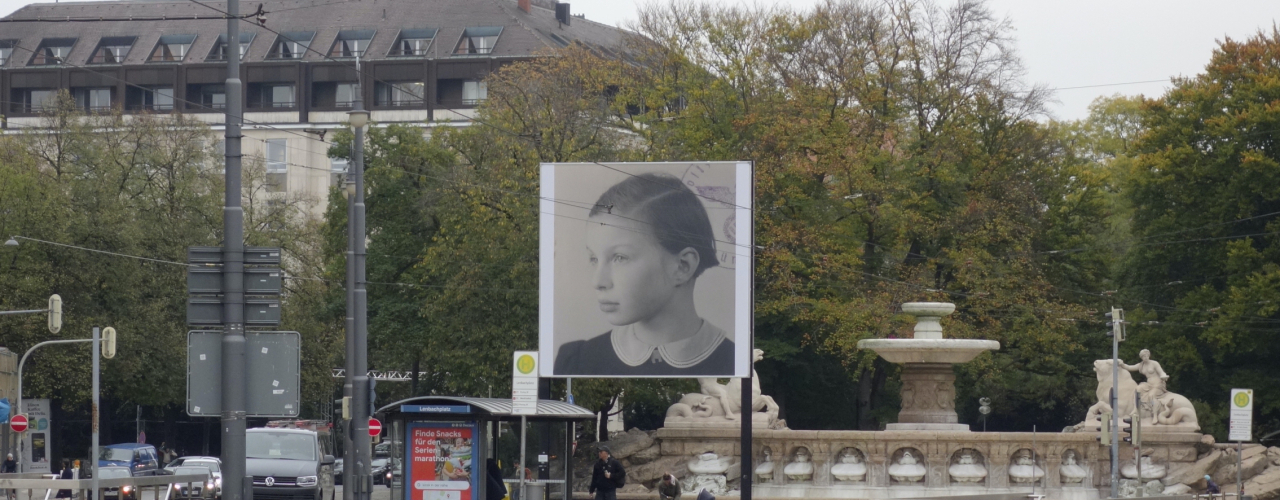 Leicht seitliche Ansicht des Billboards am Lenbachplatz. Das Motiv zeigt die Schwarz-Weiß-Fotografie eines Mädchens im Halbprofil. In der rechten Ecke sind Teile eines runden Stempelabdruckes zu sehen, im Zentrum der Reichsadler mit Hakenkreuz im Eichenkranz sowie der Schriftzug "Polizeipräsidium München".