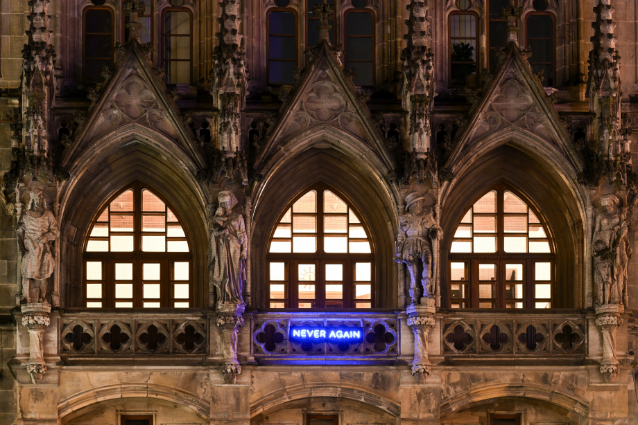 Fotografie eines Ausschnitts der Fassade des Münchner Neuen Rathauses. Am Rathausbalkon ist der Schriftzug "NEVER AGAIN" in blauleuchtenden Neonbuchstaben angebracht, ein Kunstwerk des Künstlers Jan Kuck. Die Räume dahinter sind hell erleuchtet.