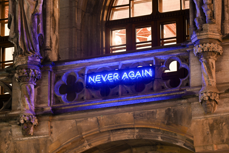 Close-Up-Fotografie eines Ausschnitts der Fassade des Münchner Neuen Rathauses. Am Rathausbalkon ist der Schriftzug "NEVER AGAIN" in blauleuchtenden Neonbuchstaben angebracht, ein Kunstwerk des Künstlers Jan Kuck. Die Räume dahinter sind hell erleuchtet.