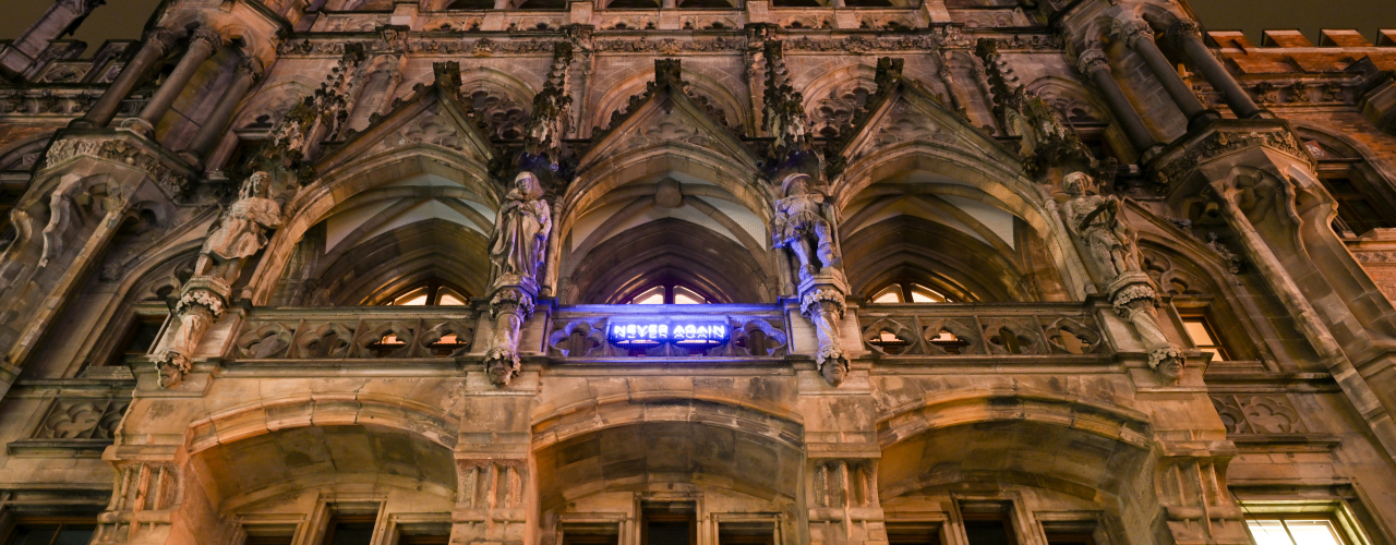 Unteransicht des Neuen Rathaus in München. An der Balkonfassade ist der Schriftzug "NEVER AGAIN" in blauleuchtenden Neonbuchstaben angebracht, ein Kunstwerk des Künstlers Jan Kuck.