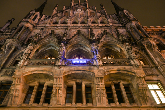 Unteransicht des Neuen Rathaus in München. An der Balkonfassade ist der Schriftzug "NEVER AGAIN" in blauleuchtenden Neonbuchstaben angebracht, ein Kunstwerk des Künstlers Jan Kuck.
