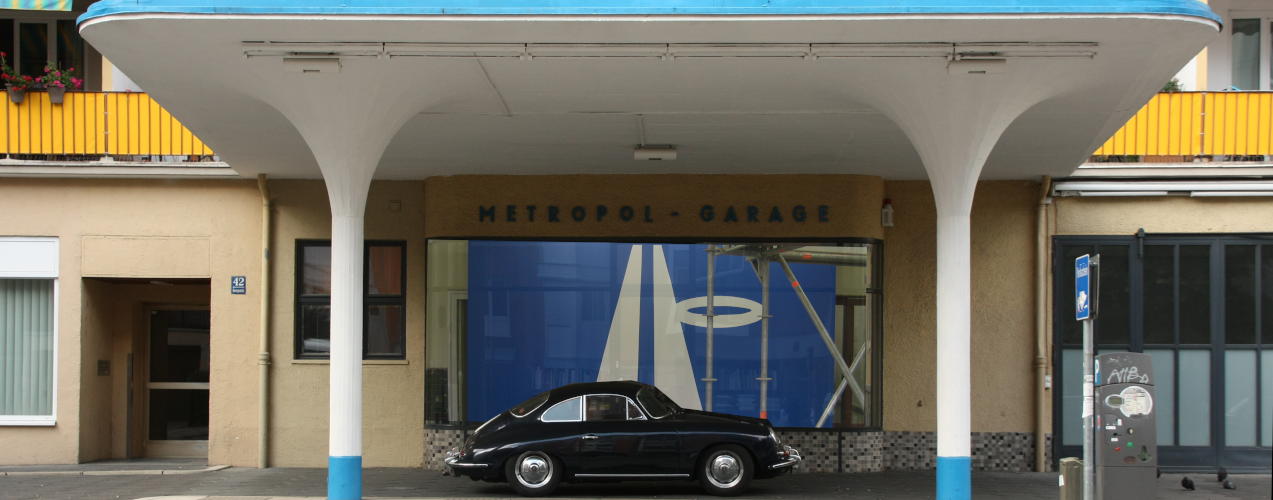 Ein schwarzer Porsche-Oldtimer ist unter dem weiß-blauen Vordach der ehemaligen Metropol Tankstelle geparkt.
