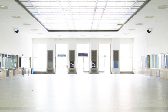 Blick in die leere Bahnhofsvorhalle des Starnberger Flügelbahnhofs mit geschlossenen Ticketschaltern. Am Kopfende der Halle, zwischen den Ausgangstüren, hängen schwarze Ankündigungsplakate mit dem Projekttitel "Applaus" in weißer Schrift auf schwarzem Grund.
