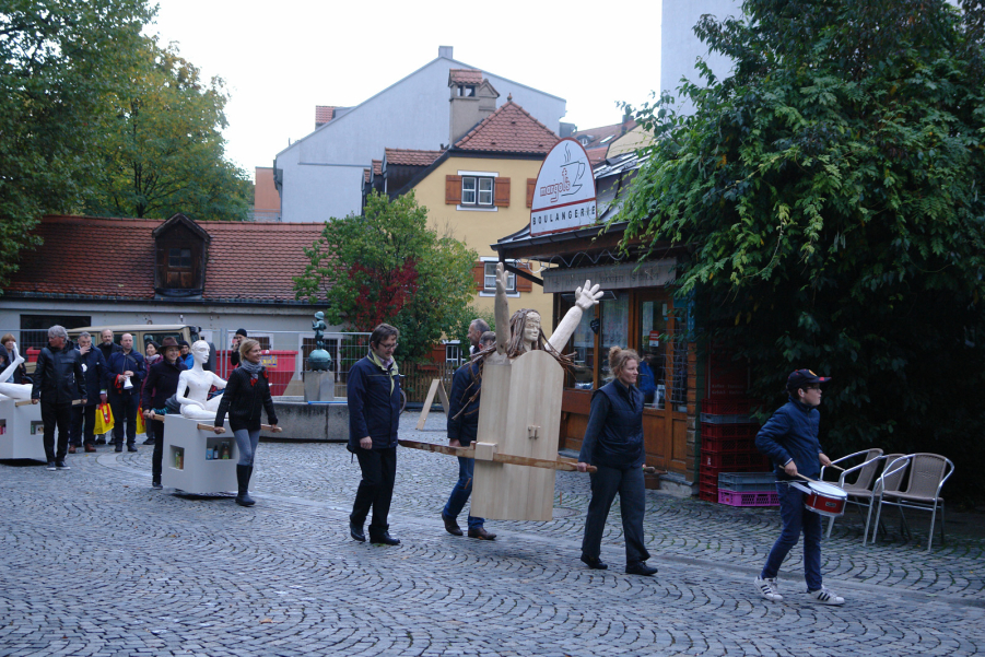 Prozession mit mehreren Menschen am Wiener Platz. Einige Teilnehmende tragen als Prozessionsgegenstände zwei Frauenfiguren, eine aus Holz und eine aus Gips, die sogenannten "Konsum-Werte-Madonnen".