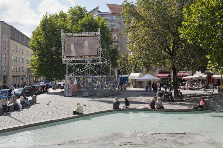 Auf dem Münchner Rindermarkt steht ein Metallgerüst mit einem aufmontierten Videoscreen, auf dem die Videoarbeit "Top View" von Nevin Aladağ gezeigt wird.