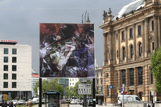 Frontale Ansicht des Billboards am Lenbachplatz stadteinwärts blickend. Das mehrfachbelichtete Foto auf dem Billboard zeigt eine Ansammlung von entsorgten Dinge in Containern. Bei längerem Hinsehen können so mehrere Schichten an unterschiedlichen Gegenständen, von Elektroschrott über Metallstangen bis hin zu Sitzpolstern, ausgemacht werden.
