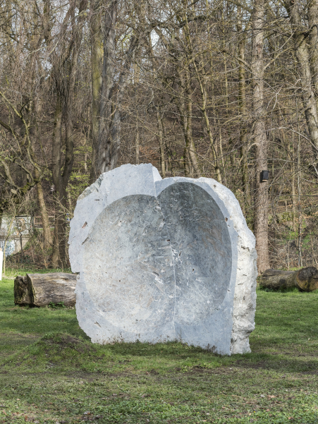 Ein großer gräulicher Stein mit konkaver Mulde steht auf Grasfläche, im Hintergrund viele Bäume