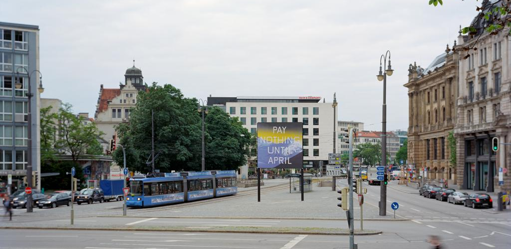 Frontale Ansicht des Lenbachplatzes mit Blick Richtung Innenstadt. In der Mitte der Platzes ist das Billboard platziert, seitlich fährt eine Trambahn dabei. Auf dem Billboard zu sehen ist eine Arbeit des Künstlers Ed Ruscha. Das Motiv zeigt eine in blau gehaltene verschneite Bergkette und darüber ein gelblicher Horizont. Auf dem Bild erscheint die Aufschrift: "Pay Nothing Until April" in weißen Großbuchstaben.