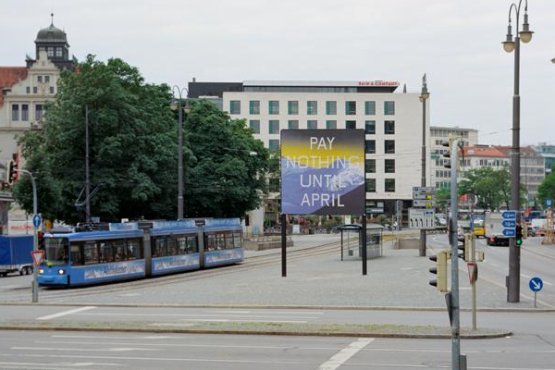 Frontale Ansicht des Lenbachplatzes mit Blick Richtung Innenstadt. In der Mitte der Platzes ist das Billboard platziert, seitlich fährt eine Trambahn dabei. Auf dem Billboard zu sehen ist eine Arbeit des Künstlers Ed Ruscha. Das Motiv zeigt eine in blau gehaltene verschneite Bergkette und darüber ein gelblicher Horizont. Auf dem Bild erscheint die Aufschrift: "Pay Nothing Until April" in weißen Großbuchstaben.