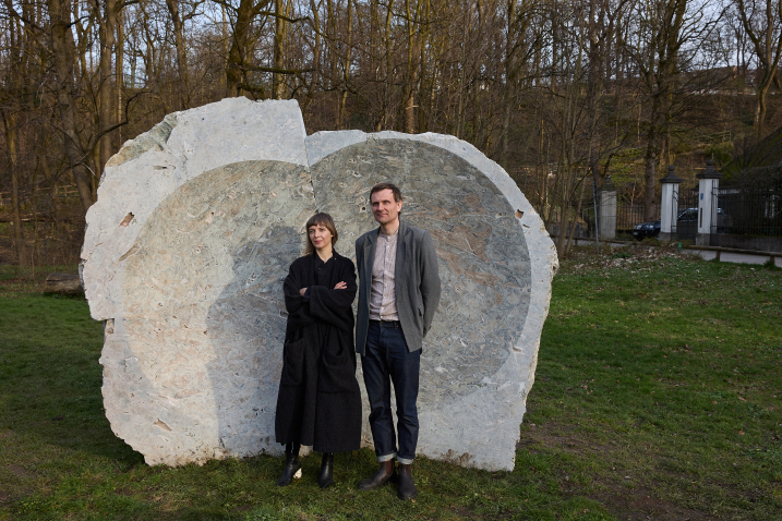 Vor einem großen gräulichen Stein mit konkaver Mulde steht eine Frau und ein Mann, Lina Lapelytė und Mantas Peteraitis, auf der Grasfläche, im Hintergrund viele Bäume