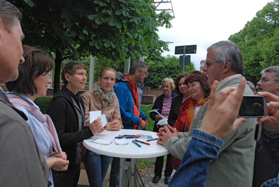Eine Gruppe von Menschen steht um einen runden weißen Stehtisch und die Gruppe befindet sich im Gespräch. Auf dem Tisch liegen Stifte und weiß-blaue Zettel. Im Hintergrund sind die Bäume des Hofgartens zu sehen.