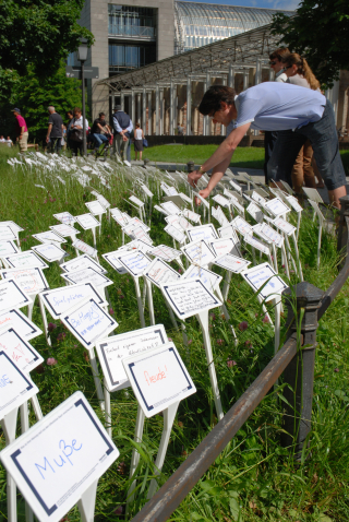 Fotografie einer Wiese im Hofgarten, in die viele weiße Schilder gesteckt sind. Auf die Schilder sind in unterschiedlichen Farben Begriffe und Sätze geschrieben. Ein Mann ist zu sehen, der noch ein Schild in die Wiese steckt. Im Hintergrund sind einige Passant*innen auf dem Spazierweg und die Bayerische Staatskanzlei zu erkennen.