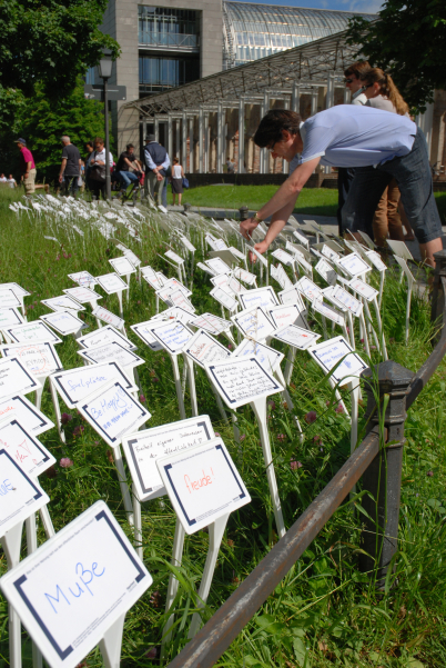 Fotografie einer Wiese im Hofgarten, in die viele weiße Schilder gesteckt sind. Auf die Schilder sind in unterschiedlichen Farben Begriffe und Sätze geschrieben. Ein Mann ist zu sehen, der noch ein Schild in die Wiese steckt. Im Hintergrund sind einige Passant*innen auf dem Spazierweg und die Bayerische Staatskanzlei zu erkennen.