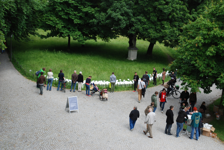 Luftaufnahme von einem Weg im Hofgarten. Zu sehen ist eine Rasenfläche, in der eine Vielzahl von weißen Schildern stecken. Vor der Rasenfläche stehen eine Reihe von Passant*innen, die die Schilder lesen. Auf dem Weg steht zudem ein Stehtisch, um den ein paar Menschen stehen.