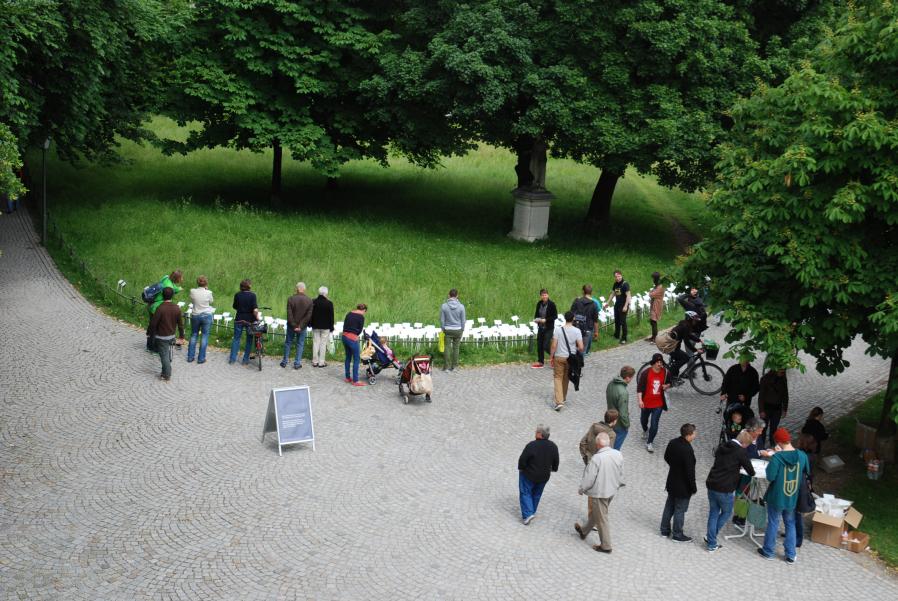 Luftaufnahme von einem Weg im Hofgarten. Zu sehen ist eine Rasenfläche, in der eine Vielzahl von weißen Schildern stecken. Vor der Rasenfläche stehen eine Reihe von Passant*innen, die die Schilder lesen. Auf dem Weg steht zudem ein Stehtisch, um den ein paar Menschen stehen.