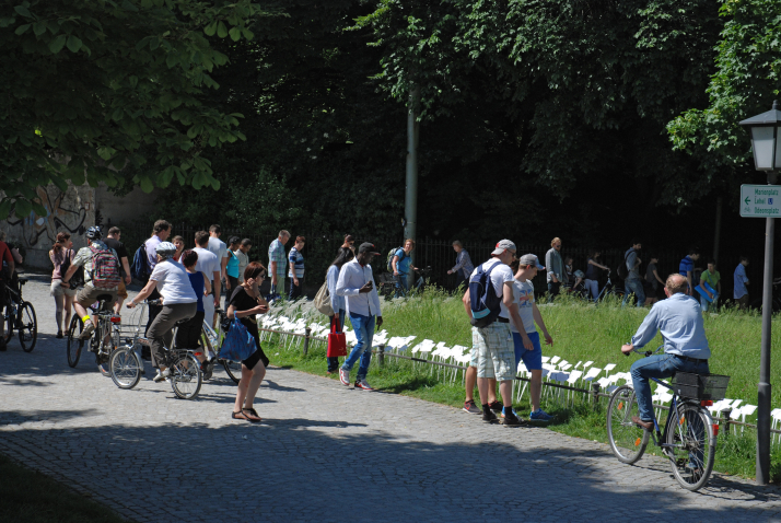 Fotografie von einem Weg im Hofgarten. Zu sehen ist eine Rasenfläche, in der eine Vielzahl von weißen Schildern stecken. Vor der Rasenfläche stehen eine Reihe von Passant*innen, die die Schilder lesen.