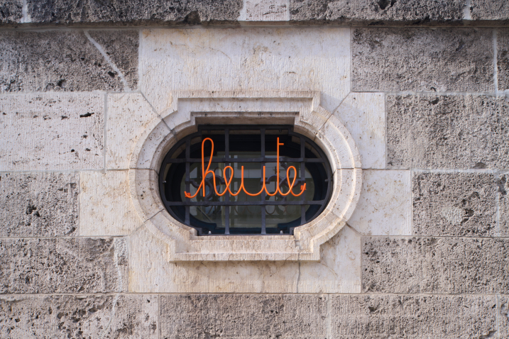 Zu sehen ist eine Hauswand mit einem Fenstergitter. In diesem Fenstergitter befindet sich ein Hakenkreuz-Muster, davor der geschwungene orangefarbene Schriftzug 'heute'.
