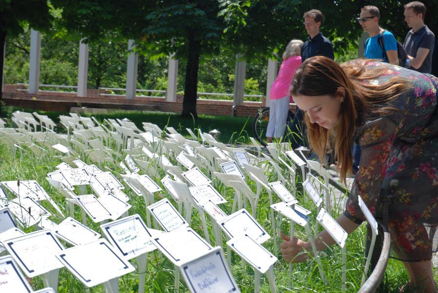 Fotografie einer Wiese im Hofgarten, in die viele weiße Schilder gesteckt sind. Auf die Schilder sind in unterschiedlichen Farben Begriffe und Sätze geschrieben. Eine Frau ist zu sehen, die noch ein Schild in die Wiese steckt. Im Hintergrund sind einige Passant*innen auf dem Spazierweg und die Bayerische Staatskanzlei zu erkennen.