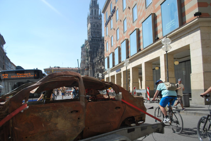 Fotografie eines braunen ausgebrannten Autowracks ohne Reifen auf dem Münchner Marienplatz vor Ludwig Beck. Das Wrack ist mit roten Spanngurten auf einen Anhänger fixiert. Im Hintergrund ist ein Stadtbus der Linie 52 zu sehen. An der Seite des Autoanhängers fahren Fahrradfahrer*innen vorbei und begutachten das ungewöhnliche Treiben.