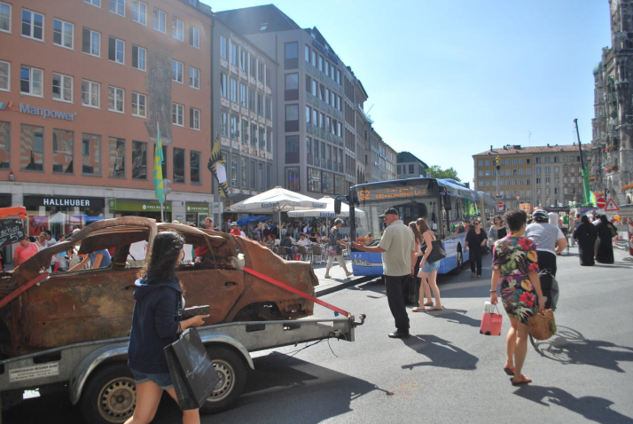 Fotografie eines braunen ausgebrannten Autowracks ohne Reifen auf dem Münchner Marienplatz. Das Wrack ist mit roten Spanngurten auf einen Anhänger fixiert. Im Hintergrund ist ein Stadtbus der Linie 52 zu sehen. Passant*innen auf dem Platz begutachten das ungewöhnliche Treiben.