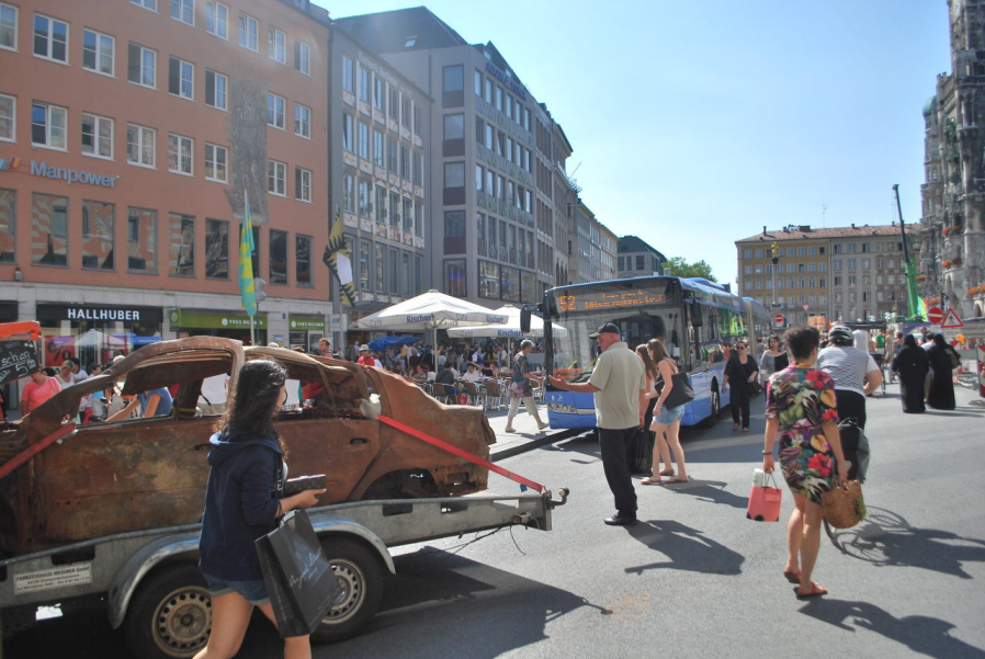 Fotografie eines braunen ausgebrannten Autowracks ohne Reifen auf dem Münchner Marienplatz. Das Wrack ist mit roten Spanngurten auf einen Anhänger fixiert. Im Hintergrund ist ein Stadtbus der Linie 52 zu sehen. Passant*innen auf dem Platz begutachten das ungewöhnliche Treiben.
