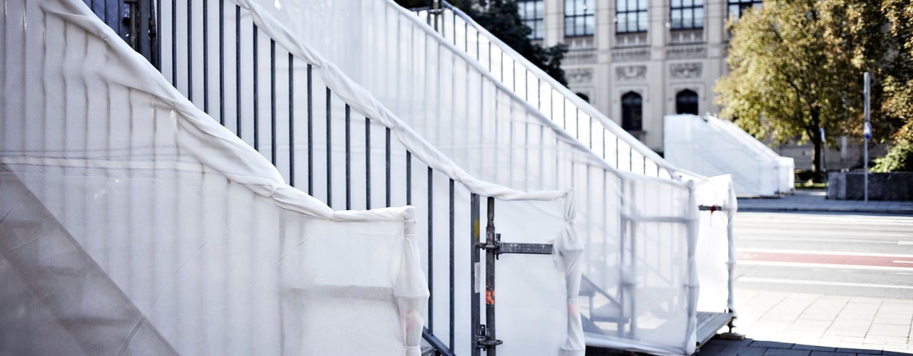 Die Fotografie zeigt zwei tribünenartige Stahltreppen, die mit weißem Stoff ummantelt sind. Die Tribünen sind auf beiden Seiten der Maximilianstraße jeweils vor den Unterführungsaufgängen, die nun zum MaximiliansForum führen, aufgestellt. Im Hintergrund zu erkennen, ist das Gebäude des Museum Fünf Kontintente, in der Bildmitte führt die Maximilianstraße entlang.