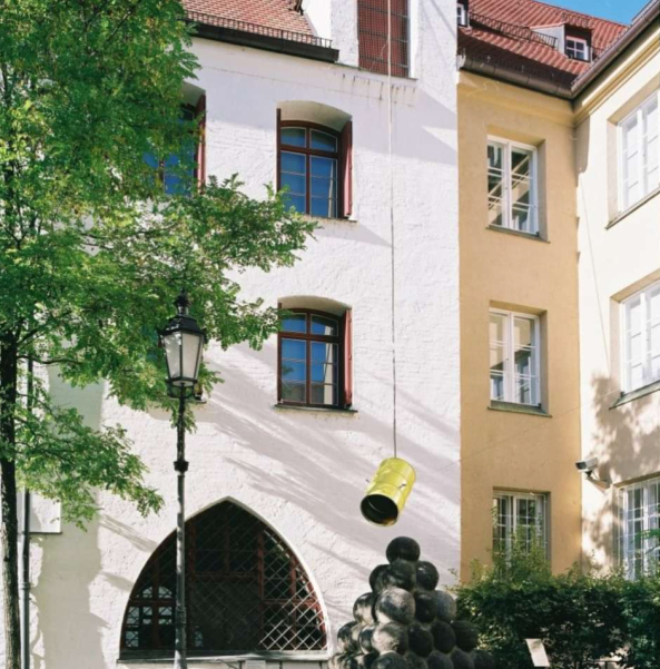 Blick in den Innenhof des Münchner Stadtmuseums. Über der Skulptur der Steinkugel-Pyramide hängt in der Luft ein gelb angemaltes Metallfass, das zwischen mehrere Seile gespannt ist. Bei dem nach unten geöffneten Fass handelt es sich um eine zeitgenössische Installation des Künstlers Florian Froese-Peeck mit dem Titel "Freisprechanlage".