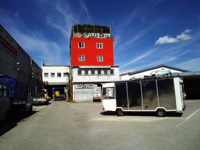 Fotografie des Grundstückes der Boxfabrik. Im Zentrum ist ein mehrstöckiges rot-graues Gebäude zu sehen, das durch zwei Schilder am Gebäude als "Boxfabrik" ausgewiesen ist. Im Hintergrund zu sehen sind einige Industriehallen. Auf dem Vorhof, vor der Boxfabrik, parkt ein weiß-schwarzer alter Kastenwagen.