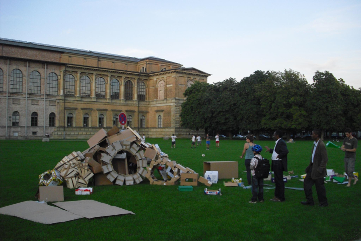 Fotografie einer Installation auf der Wiese hinter der Alten Pinakothek. Die Installation der Künstlerin Valerie Christiansen besteht aus verschiedenen alten Verpackungsmaterialien, vor allem Kartons und Obstkisten aus Sperrholz. Die unterschiedlichen Boxen sind zu Rädern und einer Art Rampe angeordnet, in der Mitte steckt ein Parkverbotsschild. Um die Installation stehen einige Passant*innen, die sie betrachten.