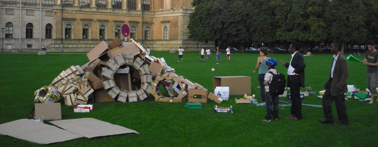 Fotografie einer Installation auf der Wiese hinter der Alten Pinakothek. Die Installation der Künstlerin Valerie Christiansen besteht aus verschiedenen alten Verpackungsmaterialien, vor allem Kartons und Obstkisten aus Sperrholz. Die unterschiedlichen Boxen sind zu Rädern und einer Art Rampe angeordnet, in der Mitte steckt ein Parkverbotsschild. Um die Installation stehen einige Passant*innen, die sie betrachten.
