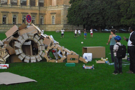 Fotografie einer Installation auf der Wiese hinter der Alten Pinakothek. Die Installation der Künstlerin Valerie Christiansen besteht aus verschiedenen alten Verpackungsmaterialien, vor allem Kartons und Obstkisten aus Sperrholz. Die unterschiedlichen Boxen sind zu Rädern und einer Art Rampe angeordnet, in der Mitte steckt ein Parkverbotsschild. Um die Installation stehen einige Passant*innen, die sie betrachten.