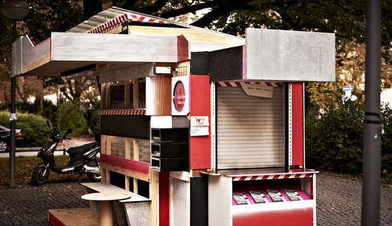Fotografie einer kioskartigen Installation, die auf einem öffentlichen Platz steht. Das in den Farben Schwarz, Rot und Weiß gehaltene überdachte Kiosk ist aus Holzplatten gefertigt und seine Seiten sind unterschiedlich gestaltet. Die Vorderseite ist mit unterschiedlichen Fächern ausgestattet, davor steht eine Bank sowie ein Tisch. Eine andere Seite hat eine Durchreiche, deren Jalousien heruntergelassen sind und darunter ein Auslagefach für Flyer.