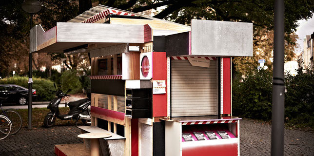 Fotografie einer kioskartigen Installation, die auf einem öffentlichen Platz steht. Das in den Farben Schwarz, Rot und Weiß gehaltene überdachte Kiosk ist aus Holzplatten gefertigt und seine Seiten sind unterschiedlich gestaltet. Die Vorderseite ist mit unterschiedlichen Fächern ausgestattet, davor steht eine Bank sowie ein Tisch. Eine andere Seite hat eine Durchreiche, deren Jalousien heruntergelassen sind und darunter ein Auslagefach für Flyer.
