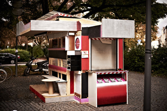 Fotografie einer kioskartigen Installation, die auf einem öffentlichen Platz steht. Das in den Farben Schwarz, Rot und Weiß gehaltene überdachte Kiosk ist aus Holzplatten gefertigt und seine Seiten sind unterschiedlich gestaltet. Die Vorderseite ist mit unterschiedlichen Fächern ausgestattet, davor steht eine Bank sowie ein Tisch. Eine andere Seite hat eine Durchreiche, deren Jalousien heruntergelassen sind und darunter ein Auslagefach für Flyer.