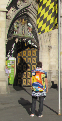 Fotografie eines Mannes, der auf dem Marienplatz vor dem Neuen Rathaus steht. Er hat den Rücken zu den Betrachtenden gewandt und steht neben einem Fahnenmast, auf dem die schwarz-gelbe-Münchenflagge gehisst ist. Er trägt eine ärmellose, bunte Weste, die offensichtlich eine Collage aus unterschiedlichen Textilstücken ist. Bei den Textilstücken handelt es sich um Textilutensilien unterschiedlicher Münchner Vereine, die zu einer patchworkartigen 'Münchner Vereinscollage' in Form eines Gewandes zusammengestellt sind.