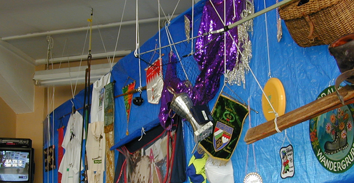 Fotografie in einen Raum, in dem eine Deckeninstallation angebracht ist. Von der Decke hängt eine große, rechteckige blaue Plastikplane. Auf dieser Plane sind unterschiedlichste Objekte angebracht, die von verschiedenen Münchner Vereinen stammen. So hängen dort u.a. verschiedene Wimpel, T-Shirts, Baseballcaps, Trophäen, Kostüme und andere Vereinsutensilien.