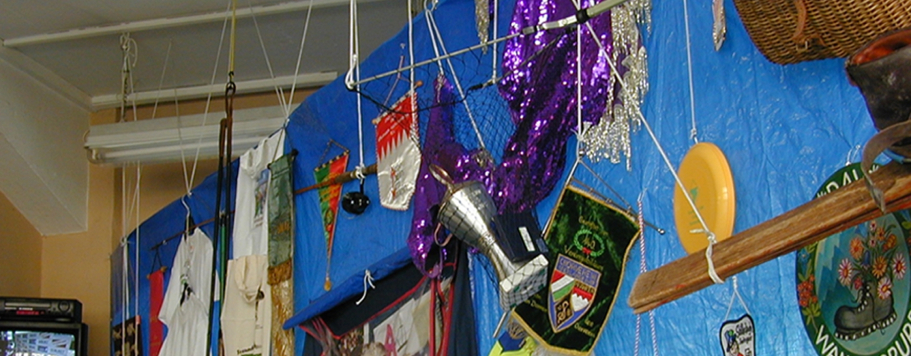 Fotografie in einen Raum, in dem eine Deckeninstallation angebracht ist. Von der Decke hängt eine große, rechteckige blaue Plastikplane. Auf dieser Plane sind unterschiedlichste Objekte angebracht, die von verschiedenen Münchner Vereinen stammen. So hängen dort u.a. verschiedene Wimpel, T-Shirts, Baseballcaps, Trophäen, Kostüme und andere Vereinsutensilien.