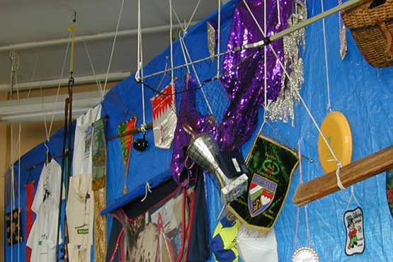 Fotografie in einen Raum, in dem eine Deckeninstallation angebracht ist. Von der Decke hängt eine große, rechteckige blaue Plastikplane. Auf dieser Plane sind unterschiedlichste Objekte angebracht, die von verschiedenen Münchner Vereinen stammen. So hängen dort u.a. verschiedene Wimpel, T-Shirts, Baseballcaps, Trophäen, Kostüme und andere Vereinsutensilien.