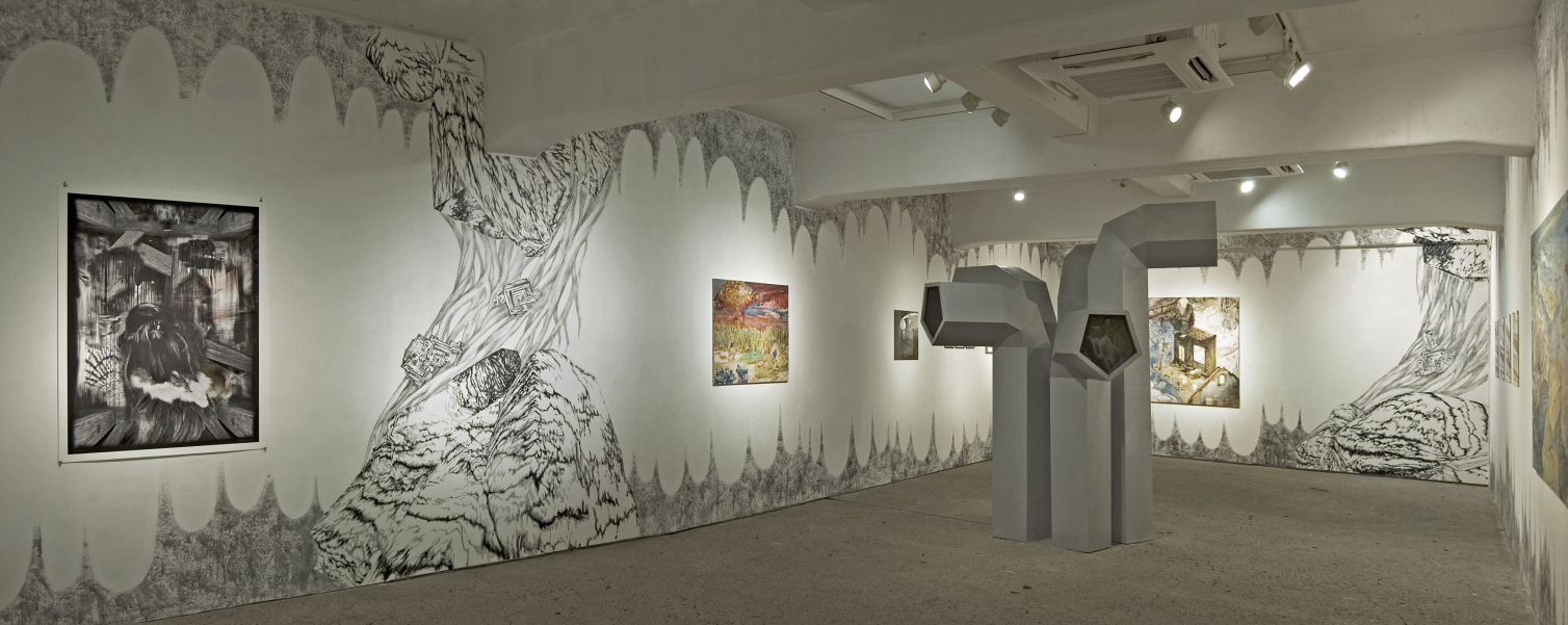 Blick in eine Ausstellung in einem fensterlosen Raum. In der Mitte des Raumes zu sehen ist eine Installation aus fünf grauen, unterschiedlich großen Objekten, die an riesige Entlüftungsrohre erinnern. An den Wänden hängen unterschiedliche Gemälde. Die Wände selbst sind auch in schwarz-weiß mit unterschiedlichen Motiven bemalt.