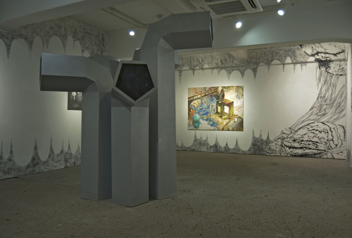 Blick in eine Ausstellung in einem fensterlosen Raum. Im Bildvordergrund zu sehen ist eine Installation aus fünf grauen, unterschiedlich großen Objekten, die an riesige Entlüftungsrohre erinnern. Im Bildhintergrund hängen zwei Gemälde an den Wänden. Die Wände selbst sind auch in schwarz-weiß mit unterschiedlichen Motiven bemalt, die zum Teil an eine abstrakte Tropfsteinhöhle erinnern.