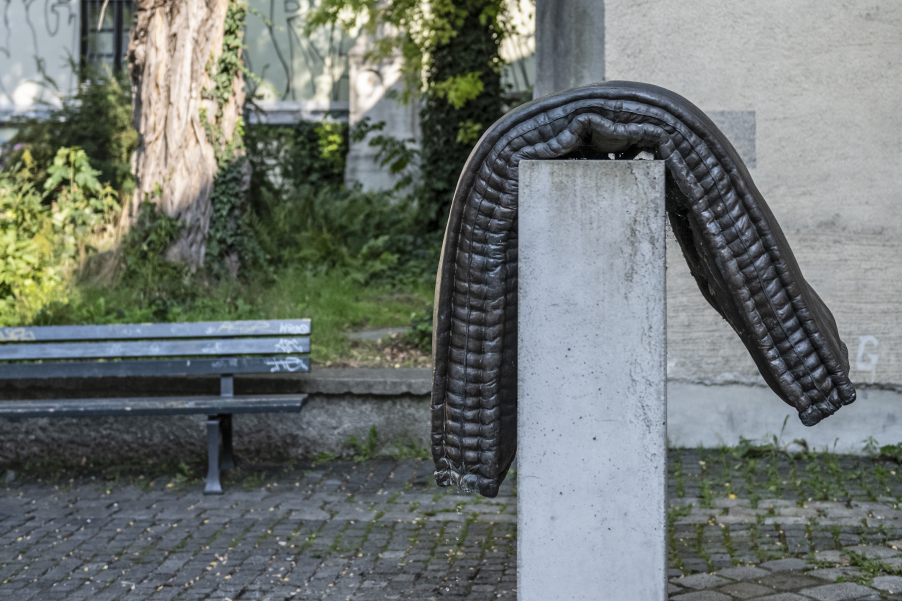 Bild einer Brunnenskulptur von Tatiana Trouvé vor der Mauer des alten Südfriedhofs am Stephansplatz. Die Skulptur besteht aus einem Brozeguss einer Matratze, die über einen Betonsockel gelegt ist.