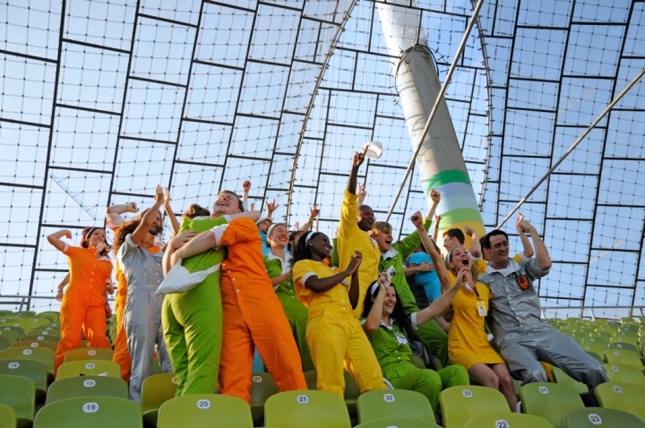 Das Bild zeigt eine im Olympiastadion aufgenommene Fotografie. In den grünen Stuhlreihen unter dem Zeltdach steht eine Gruppe von Performer*innen, die freudig und jubelnd die Arme in die Höhe strecken, in die Hände klatschen oder sich in den Armen liegen. Jede*r Performer*in trägt einen einfarbigen Overall, deren Farben von Orange und Grün zu Blau, Silber und Gelb reichen.