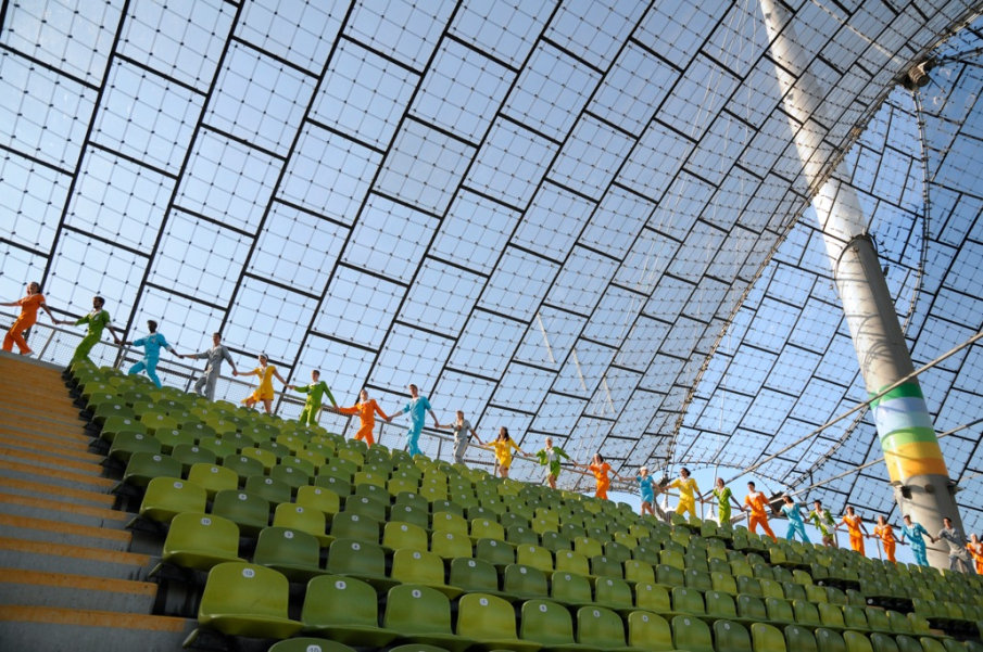 Das Bild zeigt eine im Olympiastadion aufgenommene Fotografie. Hinter der letzten grünen Stuhlreihe unter dem Zeltdach laufen Performer*innen in einer Reihe hintereinander und halten sich dabei an den Händen. Jede*r Performer*in trägt einen einfarbigen Overall, deren Farben reichen von Orange und Grün zu Blau, Silber und Gelb.