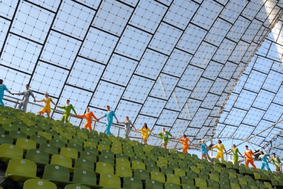 Das Bild zeigt eine im Olympiastadion aufgenommene Fotografie. Hinter der letzten grünen Stuhlreihe unter dem Zeltdach laufen Performer*innen in einer Reihe hintereinander und halten sich dabei an den Händen. Jede*r Performer*in trägt einen einfarbigen Overall, deren Farben reichen von Orange und Grün zu Blau, Silber und Gelb.