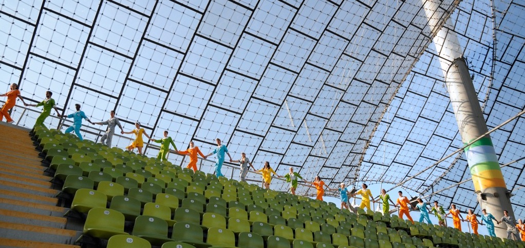 Das Bild zeigt eine im Olympiastadion aufgenommene Fotografie. Hinter der letzten grünen Stuhlreihe unter dem Zeltdach laufen Performer*innen in einer Reihe hintereinander und halten sich dabei an den Händen. Jede*r Performer*in trägt einen einfarbigen Overall, deren Farben reichen von Orange und Grün zu Blau, Silber und Gelb.