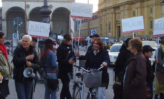 Die Fotografie zeigt eine Gruppe von Menschen, die an der Taxi-Wendeschleife beim Odeonsplatz stehen. Sie halten Megaphone und Plakate in der Hand. Auf den weißen Plakaten stehen Namen in Großbuchstaben wie "Judith", "Giehse", "Pflaum" und "Krull". Zwischen ihnen gehen und fahren Passant*innen mit Fahrrädern vorbei.